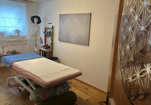 Körper Werkstatt A bright therapy room features a massage table with a pink cover and a blue towel. A human skeleton model stands nearby, and various massage tools are displayed on a wall-mounted rack. Natural light filters in through a window with sheer curtains.