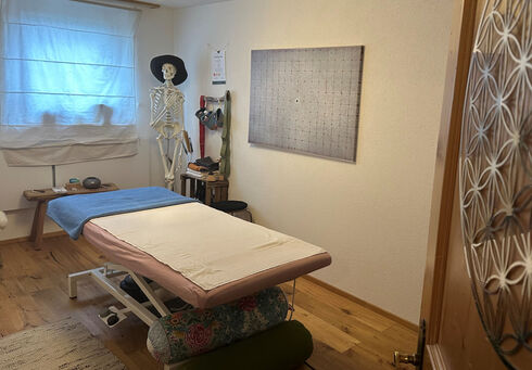Körper Werkstatt Das Bild zeigt einen Behandlungsraum mit einem Massagetisch, der mit einer blauen Decke bedeckt ist. An der Wand hängt ein großes Bild mit einem Rastermuster. Ein Skelettmodell steht neben dem Tisch, und es gibt einige therapeutische Utensilien in der Nähe.