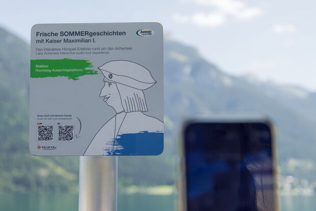 Hörspiel-Erlebnis am Achensee mit Kaiser Maximilian I. Ein Informationsschild am Aussichtspunkt Hochsteg am Achensee. Es bewirbt eine interaktive Hörspiel-Tour mit Kaiser Maximilian I. und enthält Anweisungen zum Scannen eines QR-Codes, um mehr über die Geschichte und die Umgebung zu erfahren.