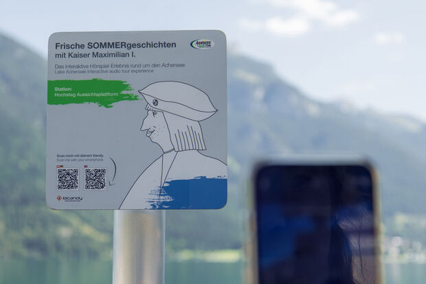 Lake Achensee interactive audio tour experience with Emperor Maximilian A sign promoting an interactive audio experience titled "Fresh Summer Stories with Emperor Maximilian I." It indicates the location as "Hochsteg Viewing Platform" and includes instructions to scan a QR code with a smartphone for more information. The background features mountains and a lake.
