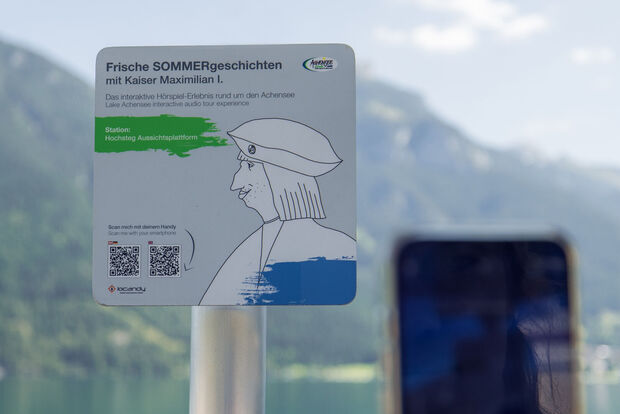 Lake Achensee interactive audio tour experience with Emperor Maximilian A sign promoting an interactive audio experience titled "Fresh Summer Stories with Emperor Maximilian I." It indicates the location as "Hochsteg Viewing Platform" and includes instructions to scan a QR code with a smartphone for more information. The background features mountains and a lake.