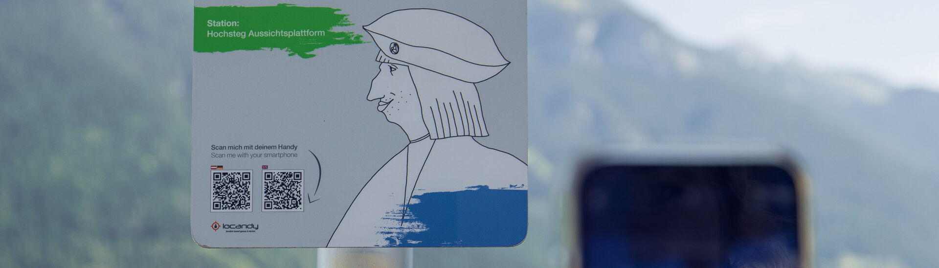 Lake Achensee interactive audio tour experience with Emperor Maximilian A sign promoting an interactive audio experience titled "Fresh Summer Stories with Emperor Maximilian I." It indicates the location as "Hochsteg Viewing Platform" and includes instructions to scan a QR code with a smartphone for more information. The background features mountains and a lake.