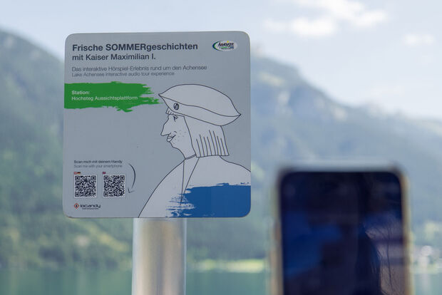 Hörspiel-Erlebnis am Achensee mit Kaiser Maximilian I. Ein Informationsschild am Aussichtspunkt Hochsteg am Achensee. Es bewirbt eine interaktive Hörspiel-Tour mit Kaiser Maximilian I. und enthält Anweisungen zum Scannen eines QR-Codes, um mehr über die Geschichte und die Umgebung zu erfahren.