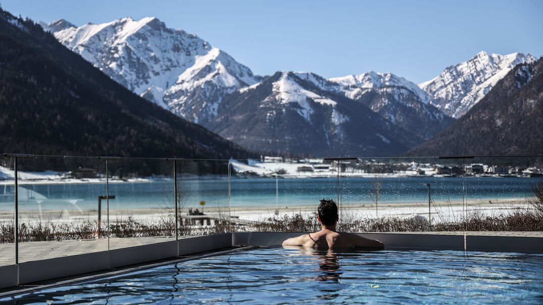 Atoll Achensee in winter This photo captures a man relaxing in the outdoor pool of the leisure centre Atoll Achensee in Maurach and enjoying the view of the lake and its surrounding mountains.