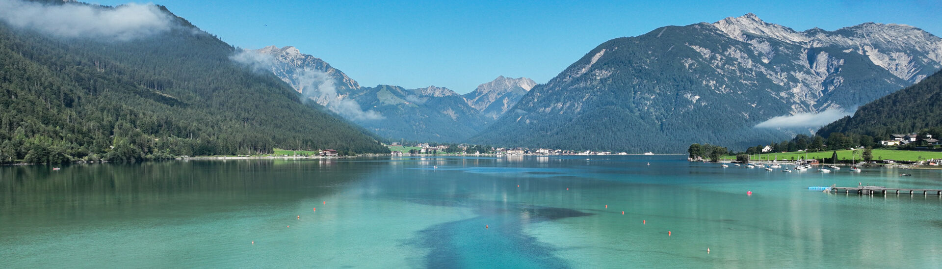 Wankratzbach on the southern shore of Lake Achensee The Lake Achensee is bordered by lush green mountains under a clear blue sky. The water displays shades of turquoise and hints of reflection from the surrounding landscape. A narrow channel leads through patches of reeds, suggesting a peaceful and natural setting.