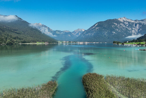 Wankratzbach on the southern shore of Lake Achensee The Lake Achensee is bordered by lush green mountains under a clear blue sky. The water displays shades of turquoise and hints of reflection from the surrounding landscape. A narrow channel leads through patches of reeds, suggesting a peaceful and natural setting.