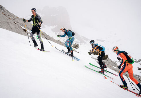 Achensee Xtreme – Individual Race Four skiers ascend a snowy slope, each using ski poles for support. They wear various brightly colored ski gear as they navigate the steep terrain. The environment is mountainous and overcast, creating a wintry scene.