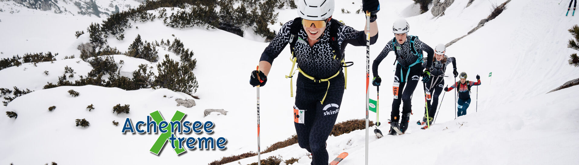 Achensee Xtreme A group of skiers ascends a snowy mountain, navigating through a winter landscape with patches of vegetation. The lead skier demonstrates determination while using ski poles, and the background shows additional skiers making their way up the slope in a cold, cloudy setting.