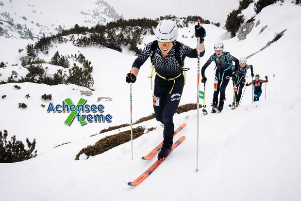 Achensee Xtreme Eine Gruppe von Skitourengehern erklimmt einen schneebedeckten Hang. Die Athleten tragen spezielle Skiausrüstung und konzentrieren sich auf den Aufstieg. Im Vordergrund ist ein Teilnehmer sichtbar, der lächelt und Stöcke zum Vorankommen nutzt. Im Hintergrund sind weitere Skifahrer zu sehen.