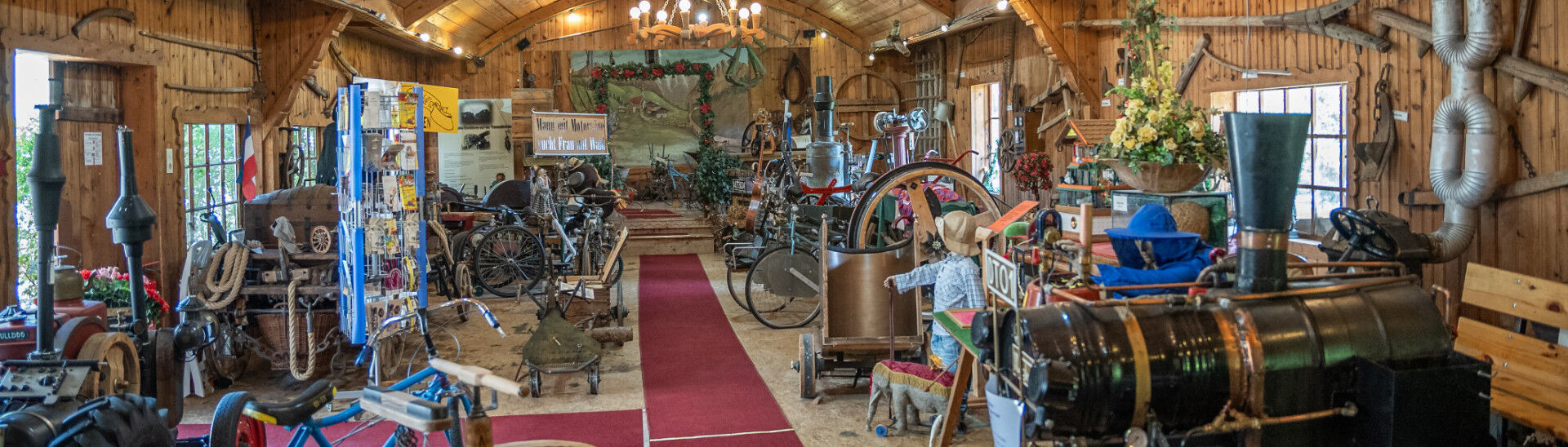 Achensee Museum World The image depicts a wide interior of a rustic museum filled with vintage machinery and artifacts. A red carpet runs down the center, flanked by various historical items, including bicycles and engines, under a wooden ceiling with warm lighting and decorations, giving a nostalgic atmosphere.