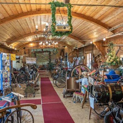 Achenseer Museums- und Erlebniswelt in Maurach am Achensee Das Bild zeigt das Innere der Achenseer Museums- und Erlebniswelt mit Holzdecke. Es sind verschiedene alte Maschinen und Werkzeuge ausgestellt, darunter Fahrräder und landwirtschaftliche Geräte. Ein roter Teppich verläuft durch den Raum, der mit Pflanzen und dekorativen Elementen geschmückt ist.