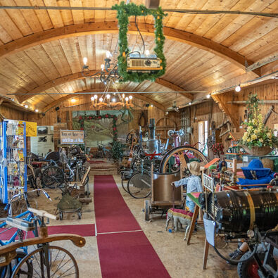 Achensee Museum World The image depicts a wide interior of a rustic museum filled with vintage machinery and artifacts. A red carpet runs down the center, flanked by various historical items, including bicycles and engines, under a wooden ceiling with warm lighting and decorations, giving a nostalgic atmosphere.