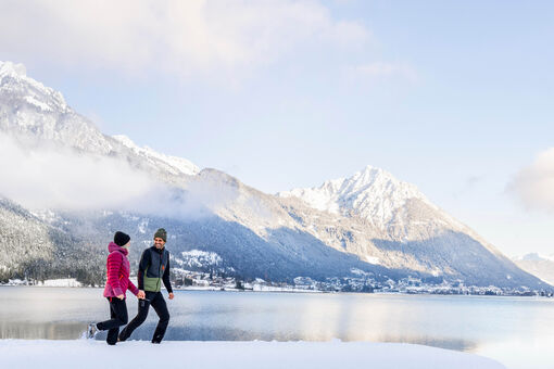 A couple enjoying a winter walk in the snowy landscape of Lake Achensee, backdropped by Maurach and the Ebner Joch.