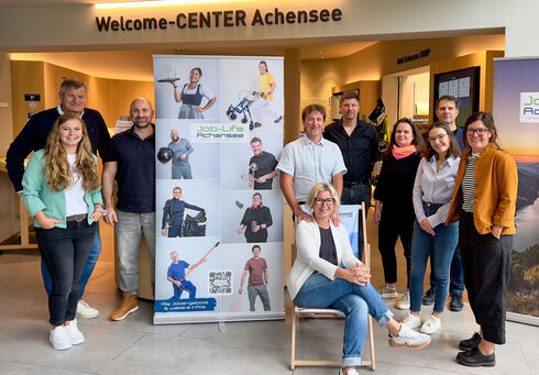 Innovative Recruiting-App Eine Gruppe von zehn Personen steht und sitzt im Welcome-CENTER Achensee im Rahmen des Leuchtturmprojekt Digitalisierung. Tourismus. vor zwei Informationsbannern. Sie lächeln freundlich.