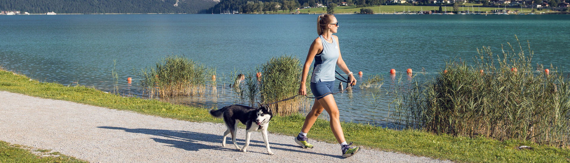 Dogs at Lake Achensee A woman walks alongside lake Achensee on a sunny day, accompanied by a black and white dog. The surrounding landscape features mountains and greenery, creating a peaceful outdoor scene. The path is gravel, bordered by grassy areas and gentle water.