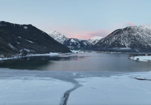 Eisbaden und Eisschwimmen im Achensee Der Blick zeigt die ruhige Winterlandschaft am Südufer des Achensees mit einem teilweise zugefrorenen See, umgeben von schneebedeckten Bergen. Der Himmel ist blau mit zarten rosa Wolken.