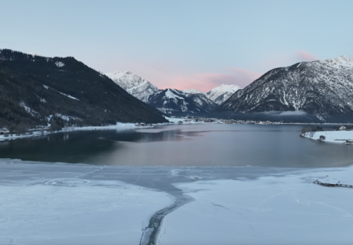 Eisbaden und Eisschwimmen im Achensee Der Blick zeigt die ruhige Winterlandschaft am Südufer des Achensees mit einem teilweise zugefrorenen See, umgeben von schneebedeckten Bergen. Der Himmel ist blau mit zarten rosa Wolken.