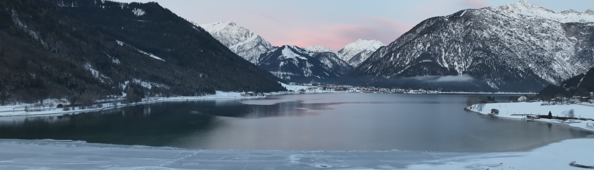 Das Südufer des Achensees im Winter Der Blick zeigt die ruhige Winterlandschaft am Südufer des Achensees mit einem teilweise zugefrorenen See, umgeben von schneebedeckten Bergen. Der Himmel ist blau mit zarten rosa Wolken.