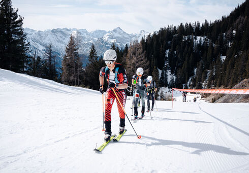 Rofan Ascent – Vertical Race A group of skiers ascends a snowy slope, navigating through the winter landscape. The background features snow-capped mountains and evergreen trees. The skiers are equipped with skiing gear and poles, concentrating on their climb. A red barrier marks the course ahead.