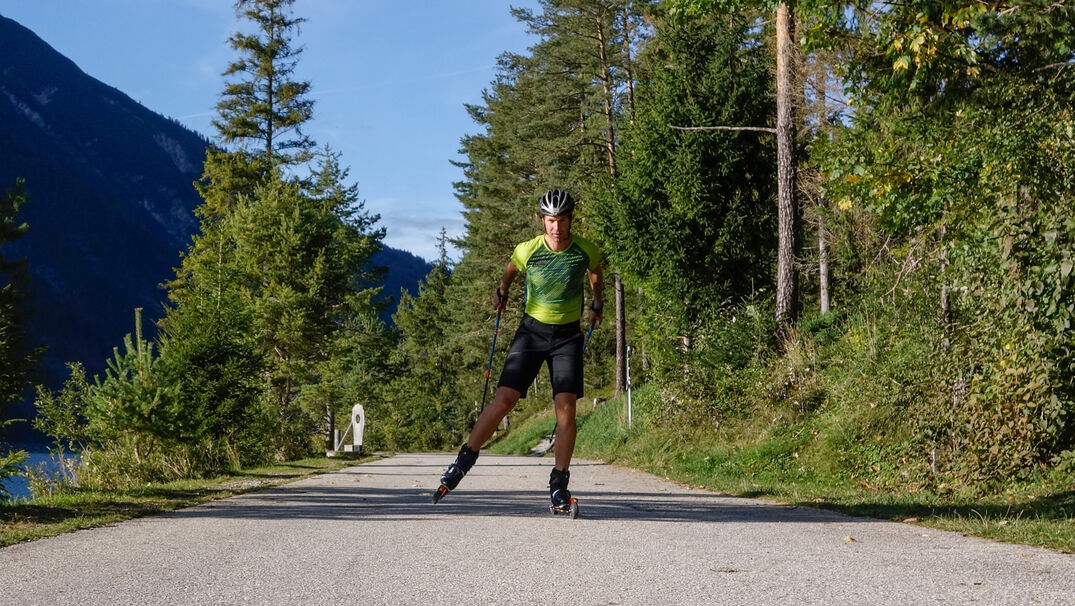 Roller skiing training at Lake Achensee A person wearing a green athletic shirt and black shorts is roller skiing on a paved path at Lake Achensee surrounded by trees and mountains.