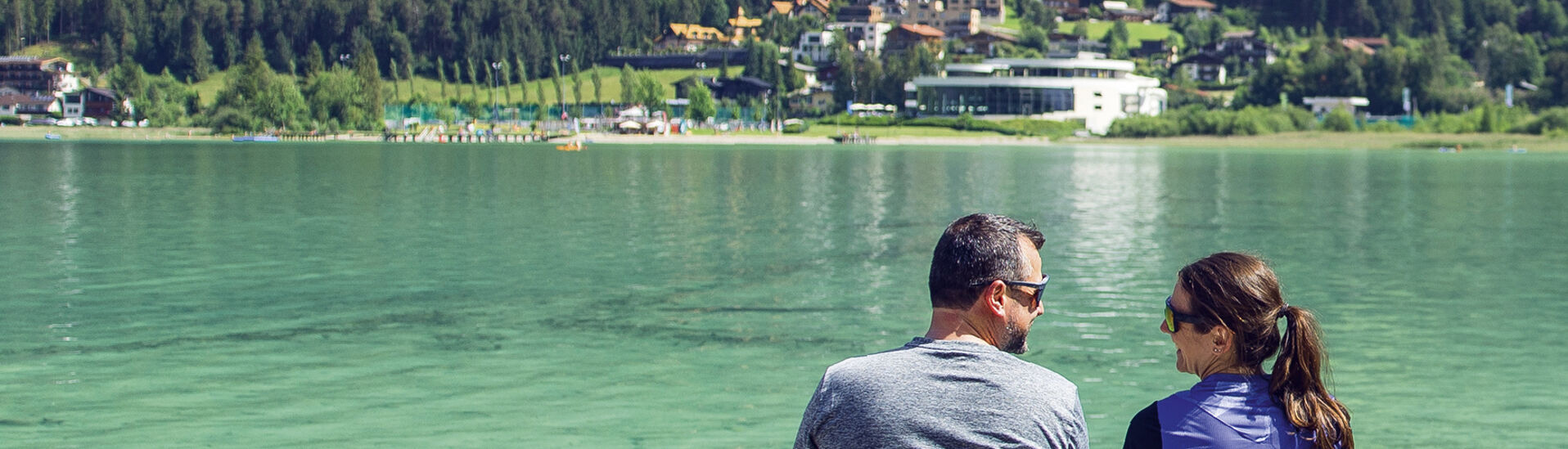 Break at the wooden footbridge in Pertisau am Achensee A man and a woman sit closely together by Lake Achensee, surrounded by lush green mountains and a picturesque village. They share a moment of companionship, gazing at the serene landscape, with reflections shimmering on the water's surface.