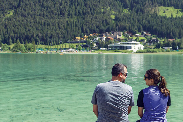Break at the wooden footbridge in Pertisau am Achensee A man and a woman sit closely together by Lake Achensee, surrounded by lush green mountains and a picturesque village. They share a moment of companionship, gazing at the serene landscape, with reflections shimmering on the water's surface.