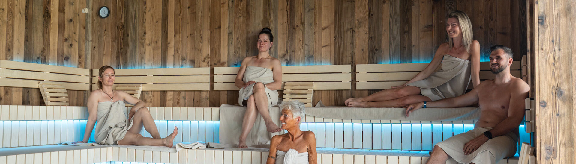 Penthouse-SPA im Atoll Achensee In einer modernen Sauna des Penthouse-SPAs im Atoll Achensee sitzen fünf Menschen in Handtüchern auf Holzbanklinien. Der Raum hat eine warme Atmosphäre mit gedämpftem Licht. In der Mitte ist ein Steingrill sichtbar.