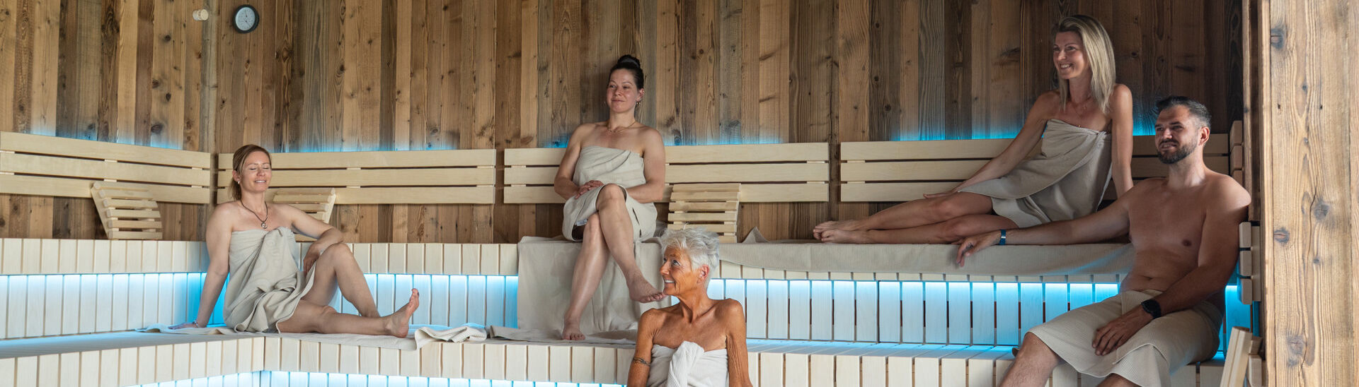 Penthouse-SPA im Atoll Achensee In einer modernen Sauna des Penthouse-SPAs im Atoll Achensee sitzen fünf Menschen in Handtüchern auf Holzbanklinien. Der Raum hat eine warme Atmosphäre mit gedämpftem Licht. In der Mitte ist ein Steingrill sichtbar.