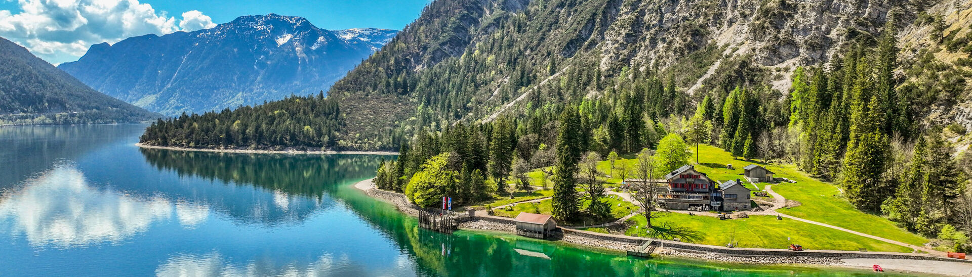 Gaisalm in der Region Achensee Der Achensee umgeben von hohen Bergen und dichtem Wald. Am Ufer steht die Gaisalm, umgeben von Wiesen. Die blau-weißen Wolken spiegeln sich im Wasser, während die Natur in frischem Grün erblüht.