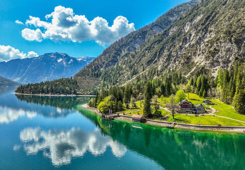 Achensee Regionsschulung Der Achensee umgeben von hohen Bergen und dichtem Wald. Am Ufer steht die Gaisalm, umgeben von Wiesen. Die blau-weißen Wolken spiegeln sich im Wasser, während die Natur in frischem Grün erblüht.