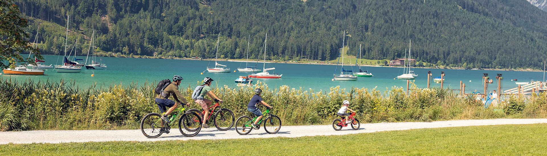 Biken mit der Familie in Maurach am Achensee Einen Familienausflug mit dem Rad am Seeufer in Maurach am Achensee machen. Im Hintergrund sind zahlreiche Segelboote zu sehen.
