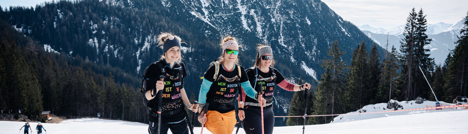 Achensee Xtreme Skitourenspaß Drei Frauen in Skibekleidung stehen auf einer präparierten Piste im Rofangebirge. Sie lächeln und halten Skistöcke. Im Hintergrund sind majestätische Berge und Bäume sichtbar. Die Stimmung ist fröhlich und sportlich, ideal für einen Skiausflug.