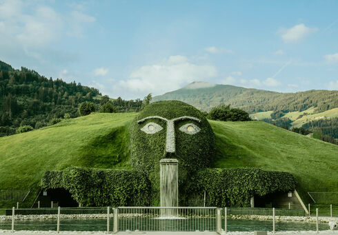 Swarovski Kristallwelten celebrate the magic of crystal The Giant of the Swarovski Kristallwelten, a large, stylized face made of greenery is situated on a grassy hill, with water flowing from its mouth into a small pond below. The background features rolling hills and trees, under a clear sky.